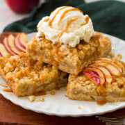 Three apple pie crumb bars stacked on a plate with a scoop of vanilla ice cream on top and drizzled with caramel sauce.