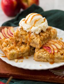 Three apple pie crumb bars stacked on a plate with a scoop of vanilla ice cream on top and drizzled with caramel sauce.