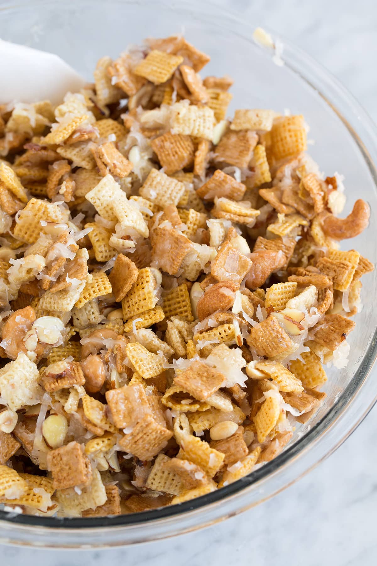 Sweet and sticky chex mix in a glass mixing bowl set over a marble surface. It includes corn and rice chex cereal, golden grahams, nuts, coconut and a butter sugar topping.