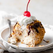 Fried ice cream cut into to show ice cream center. It is drizzled with chocolate sauce, topped with whipped cream and a maraschino cherry.