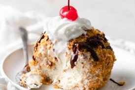 Fried ice cream cut into to show ice cream center. It is drizzled with chocolate sauce, topped with whipped cream and a maraschino cherry.