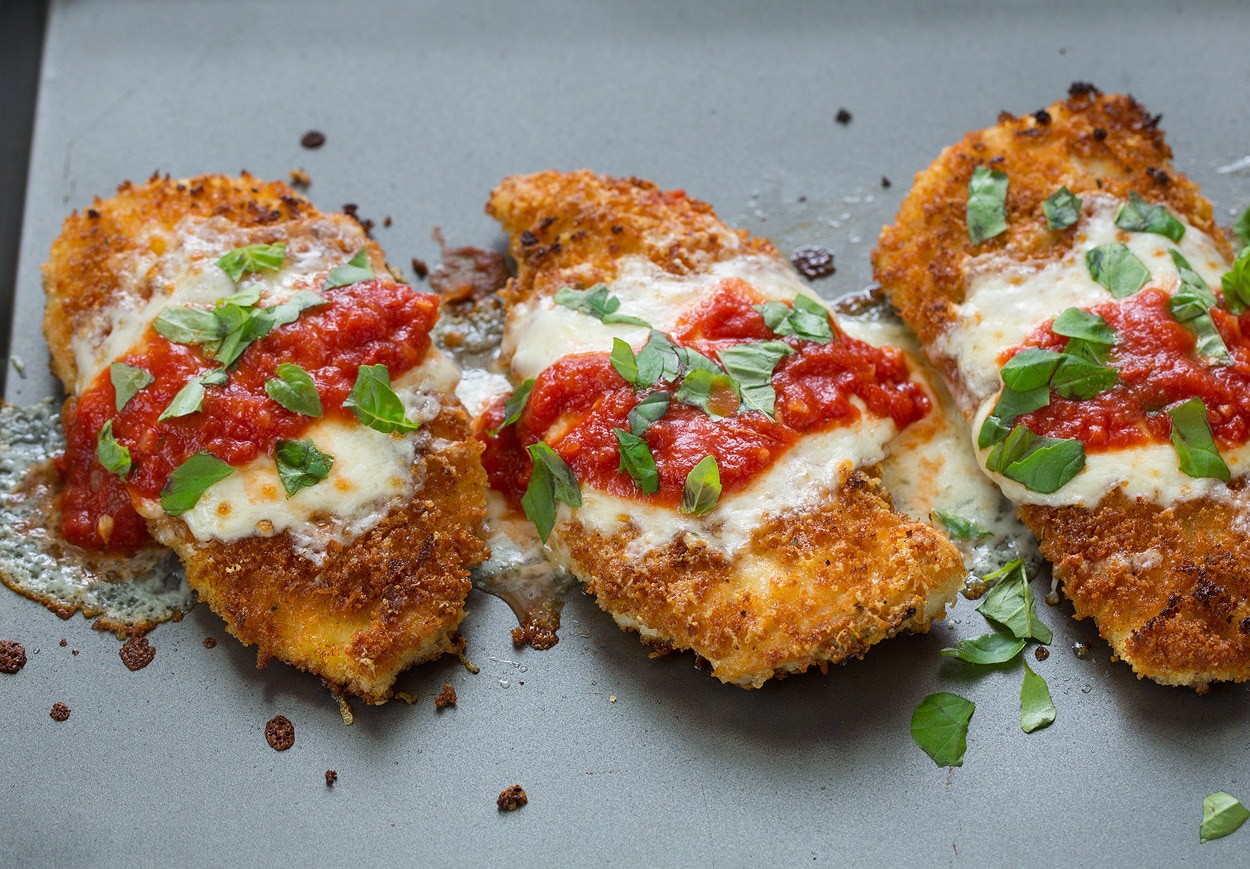 BEST Chicken Parmesan recipe! Finished chicken parmesan with cheese marinara and basil after baking. Shown on dark baking sheet.