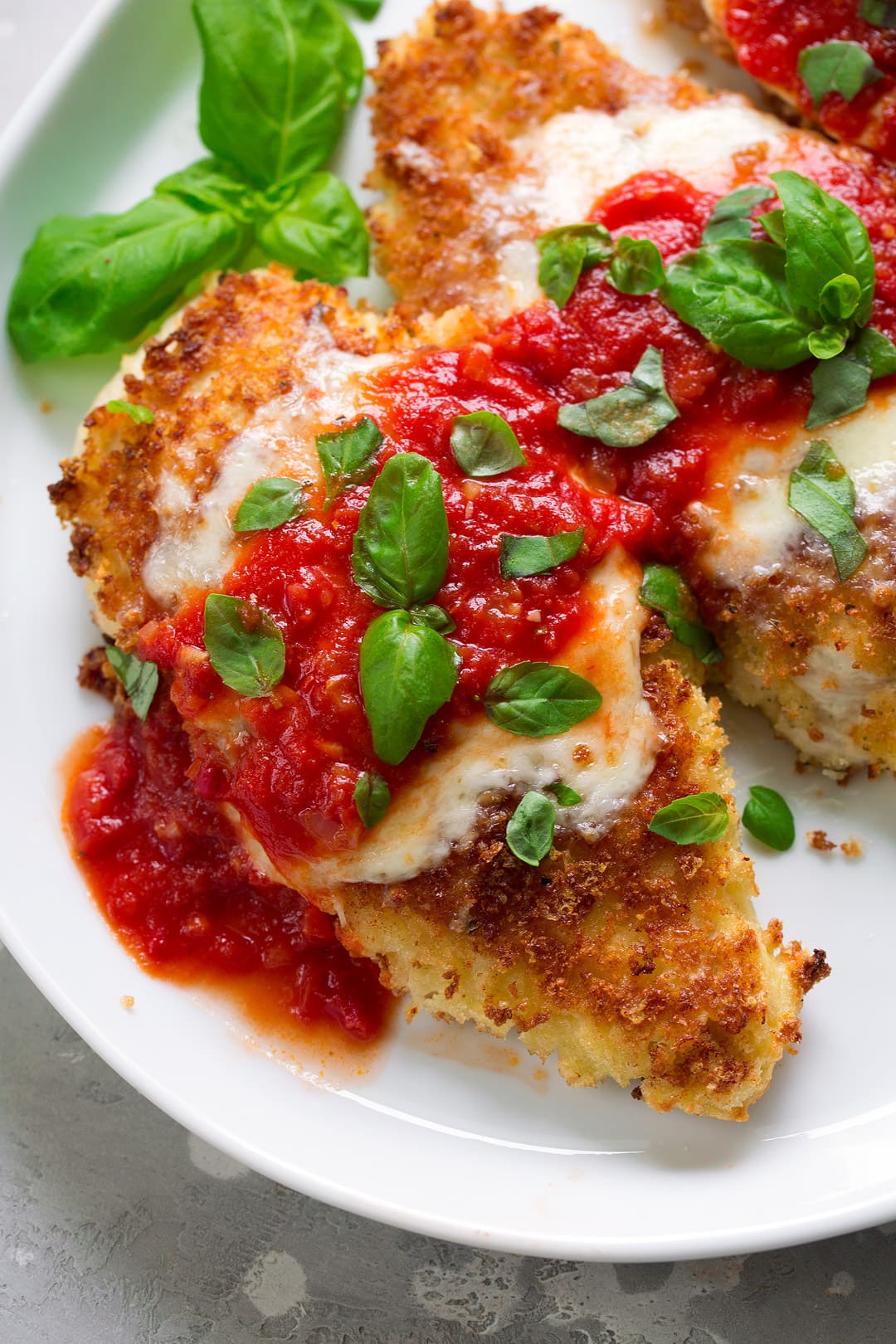 BEST Chicken Parmesan recipe! Overhead close up image of two pieces chicken parmesan on a white serving plate. Chicken is covered with golden brown Panko bread crumbs, melted mozzarella, marinara sauce and fresh basil.