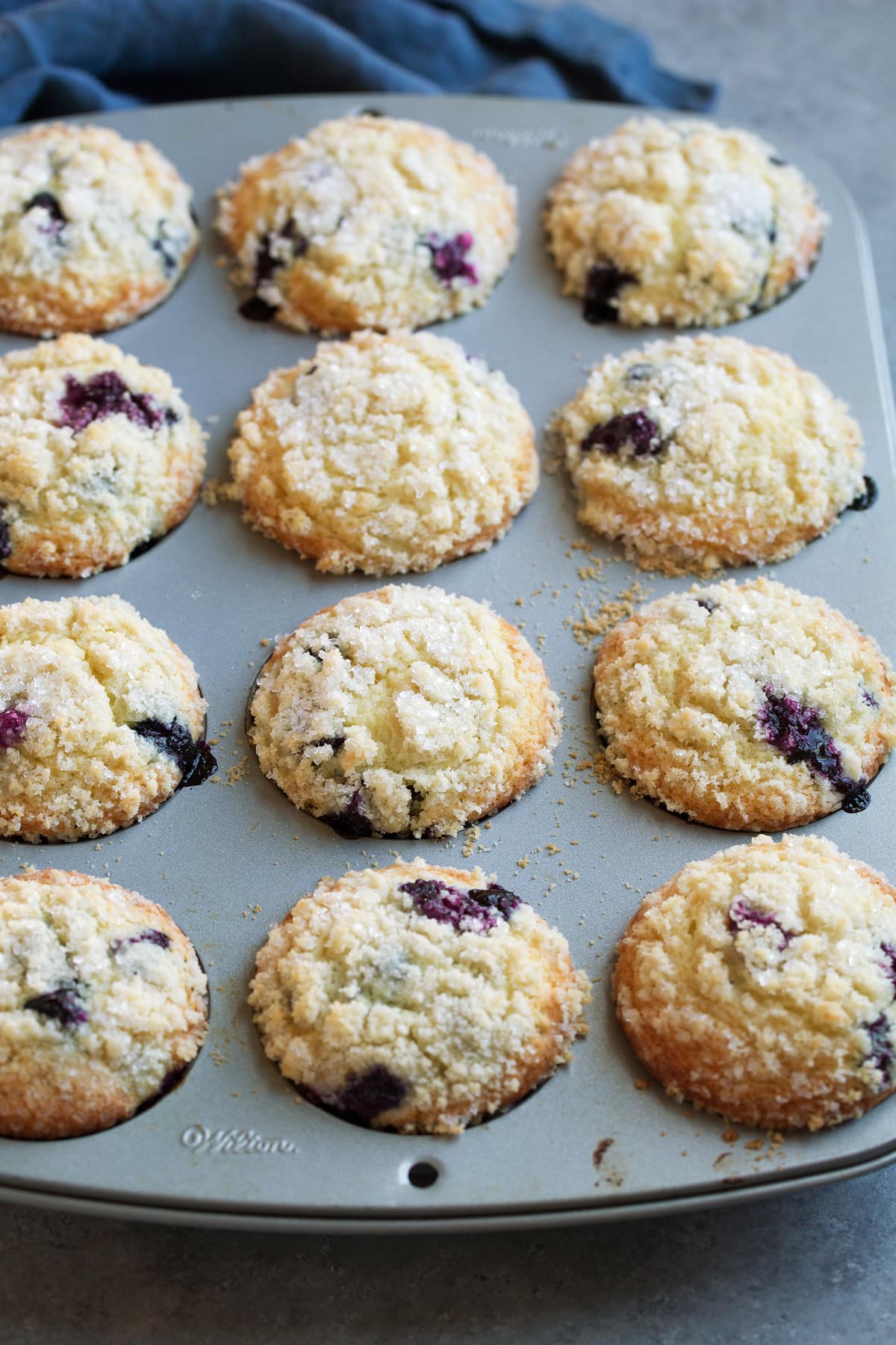 Blueberry muffins with crumb topping after baking, shown in a 12 hole muffin pan.