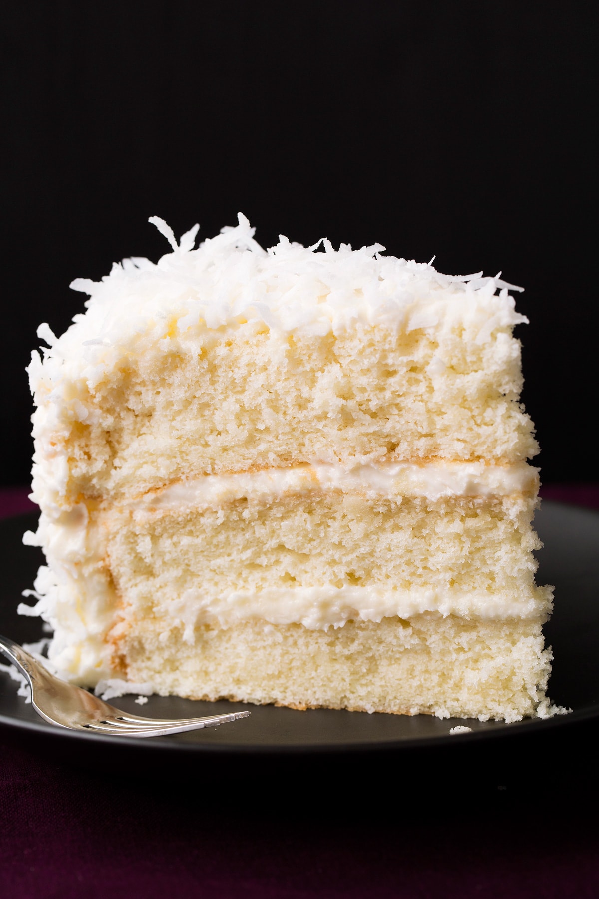 Coconut Cake Recipe Slice of three layered homemade coconut cake on a black plate with a black background.