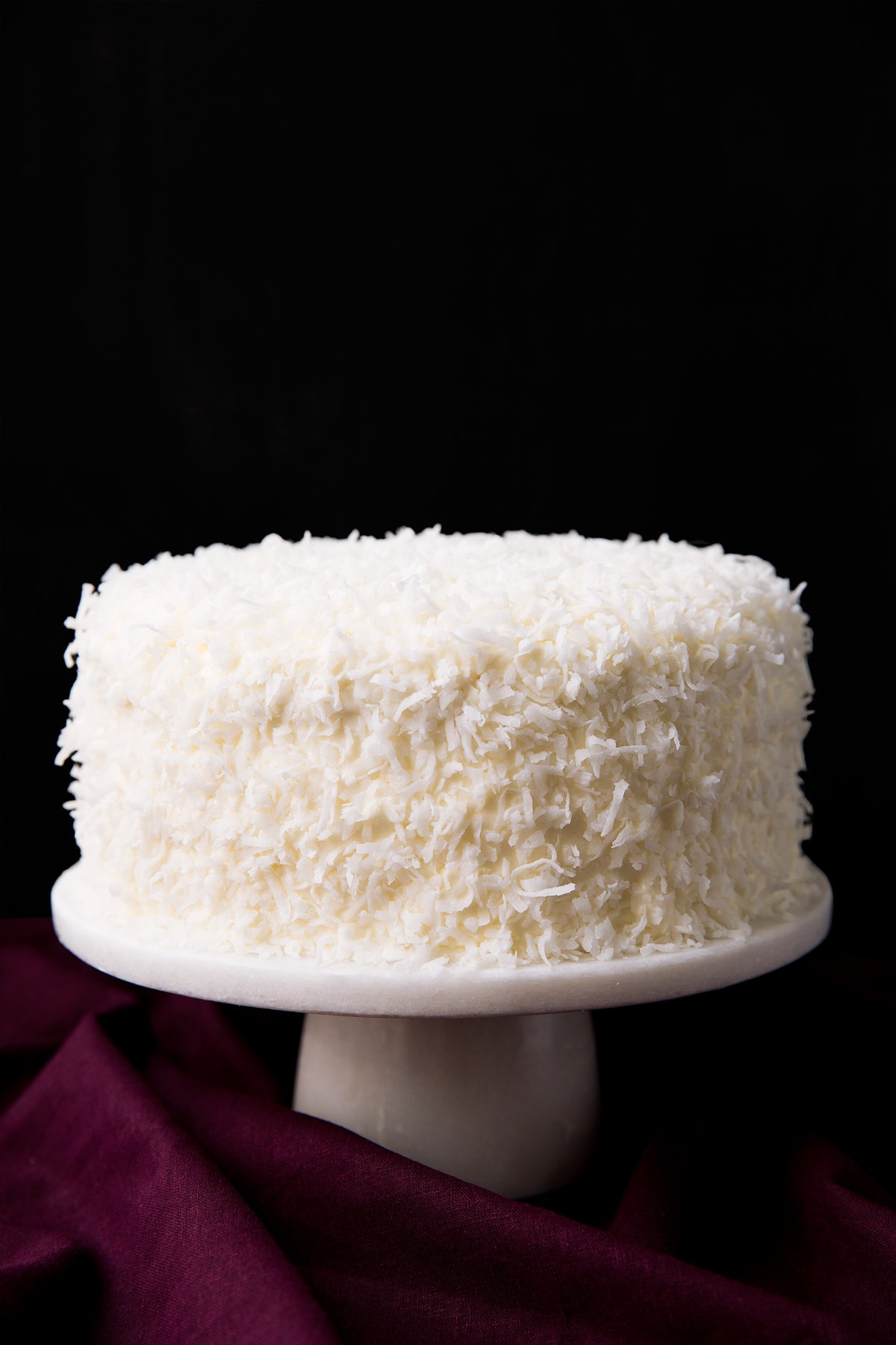Best Ever Coconut Cake! Whole made from scratch coconut cake garnished with lots of coconut and set over a white marble cake stand with a black ground and a maroon tablecloth underneath.