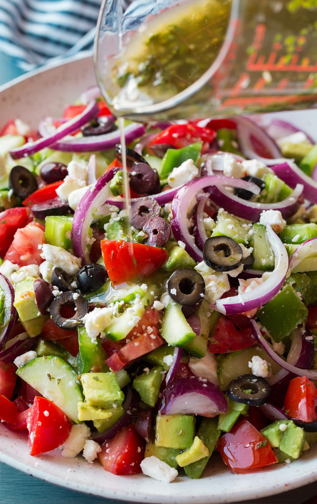 Greek Salad - this is the BEST Greek Salad! So easy to make, it's packed with veggies (love the addition of avocado), and the dressing is so flavorful. A must try recipe! #greek #salad #recipe #vegetables #sidedish #summer Greek Salad in a bowl with dressing getting poured over