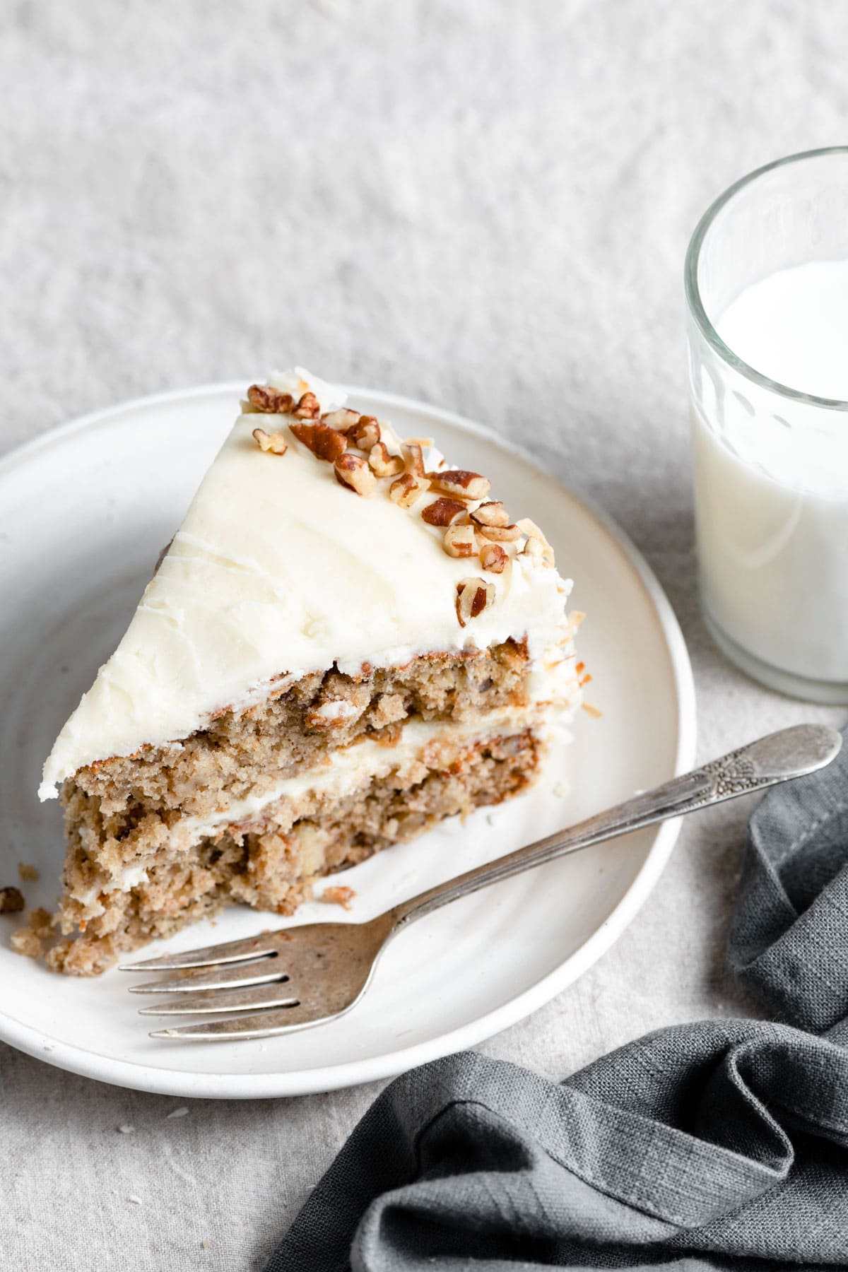 Hummingbird Cake Slice of Hummingbird Cake on a white dessert plate with a fork. Next to they plate there is a dark grey napkin and glass of milk in the background.