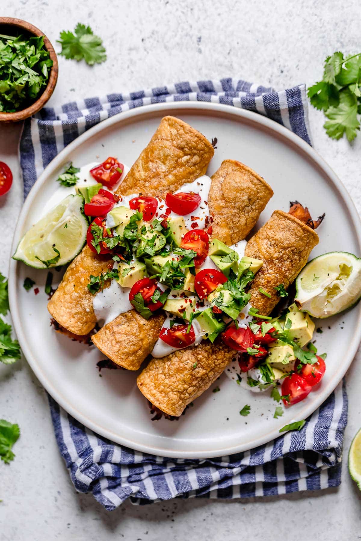Baked Chicken Taquitos Taquitos on a white plate topped with sour cream, tomatoes, avocados and cilantro. Lime wedges are set of to the side and plate is set on a blue striped napkin.