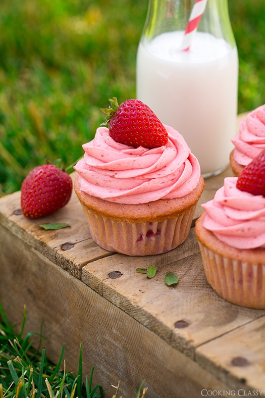 These Strawberry Cupcakes are made with chunks of fresh strawberries in the cake batter and are topped with a luscious pink Strawberry Buttercream Frosting. Bursting with flavor, these cupcakes would go down a treat at any celebration! #cookingclassy #cupcakes #strawberry #buttercream #cakes Strawberry Cupcakes on a wooden surface with a glass of milk