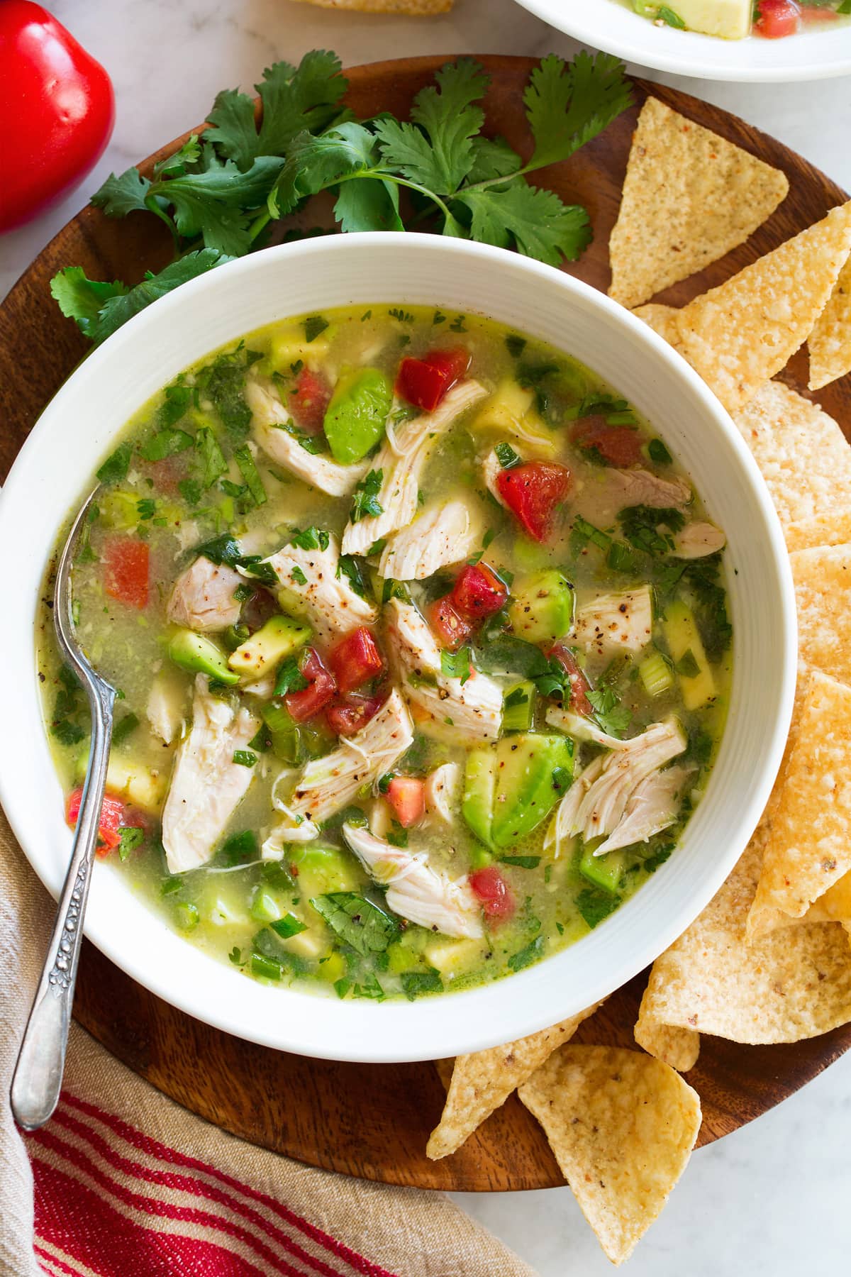 Chicken Avocado Soup Chicken and avocado soup in a serving bowl set over a wooden plate. Shown overhead with a side of tortilla chips and cilantro.
