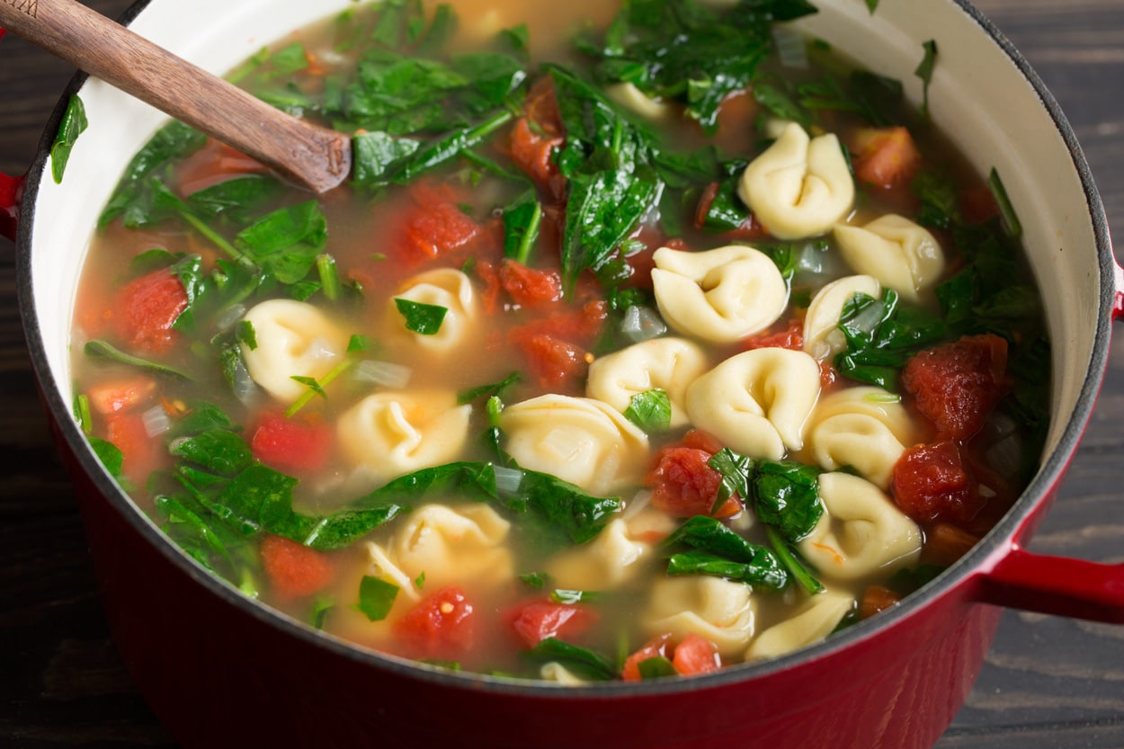 Tortellini Soup with Fresh Spinach Tomato and Garlic Completed tortellini soup in a large red cast iron pot with a wooden spoon.
