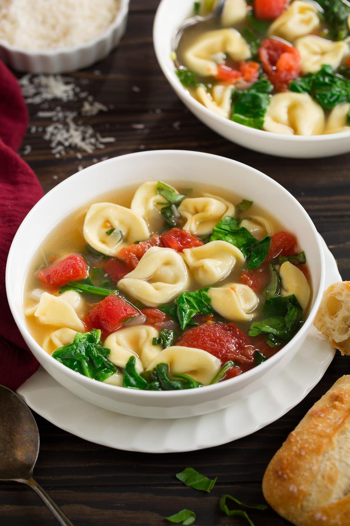Tortellini Soup with Fresh Spinach Tomato and Garlic Tortellini Soup with Spinach Tomato and Garlic in a white serving bowl set over a ruffled white plate, sitting on a dark wooden tabletop with a side of bread.