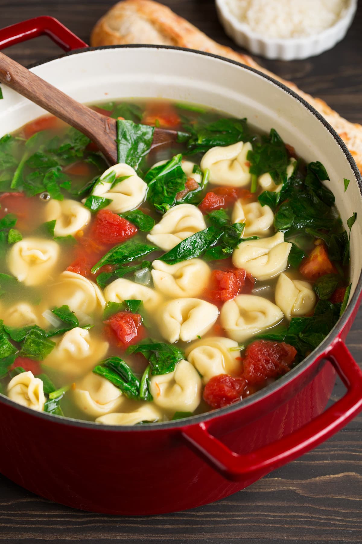 Tortellini Soup with Fresh Spinach Tomato and Garlic Close up image of tortellini soup in a red pot set over a wooden tabletop.