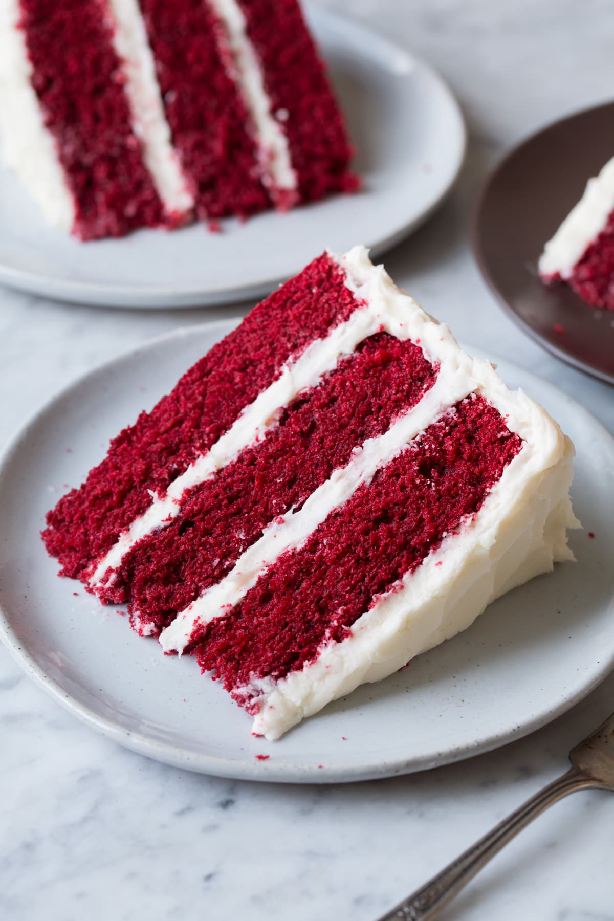 Best Ever Red Velvet Cake! Soft, moist, fluffy and perfectly rich. Single slice on a serving plate with two second slices in the background.