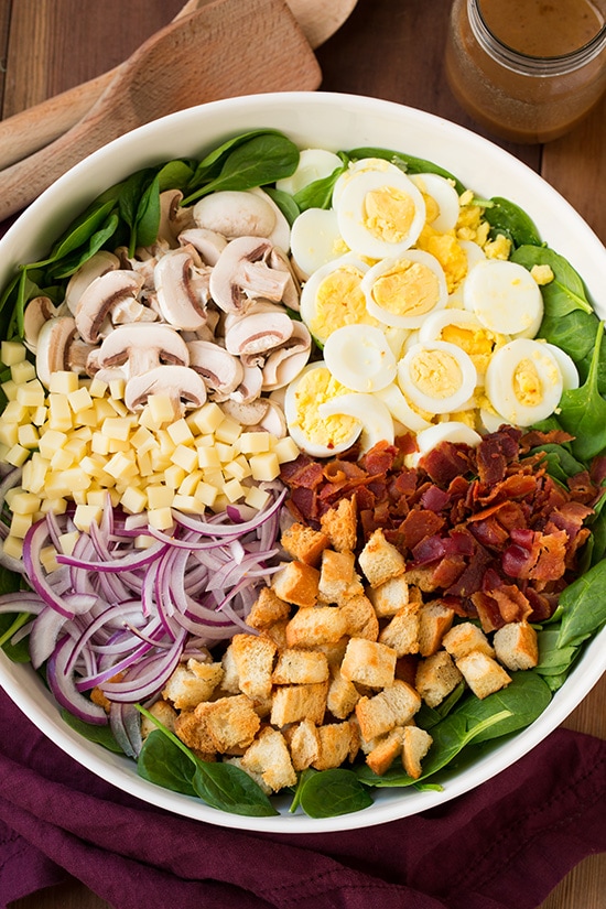 spinach salad with warm bacon dressing topped with cheese, eggs, mushrooms, bacon, croutons, and onions