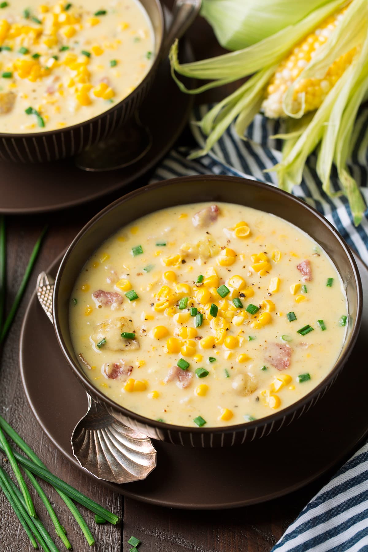 This summer Corn Chowder is creamy, comforting and full of flavor! Made with an abundance of fresh sweet corn, tender potatoes, flavorful hearty bacon, and finished with chives and cheese. Truly this soup is too hard to resist and can be enjoyed all summer long! Corn Chowder in two serving bowls. Chowder has fresh pieces of corn, chunks of ham and is garnished with chives.