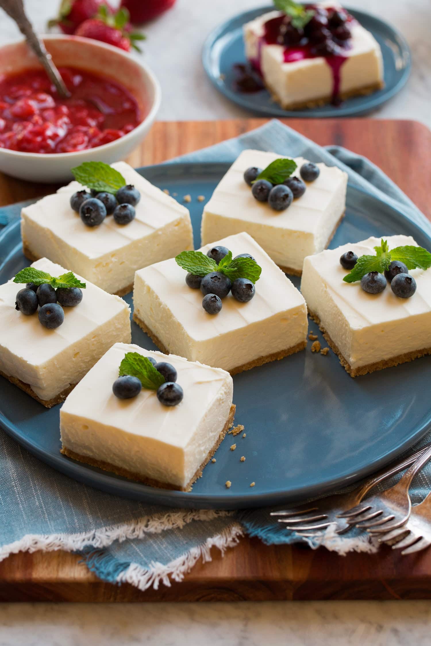 No Bake Cheesecake No bake cheesecake shown cut into bars garnished with fresh blueberries and mint. They are resting on a large blue serving plate.