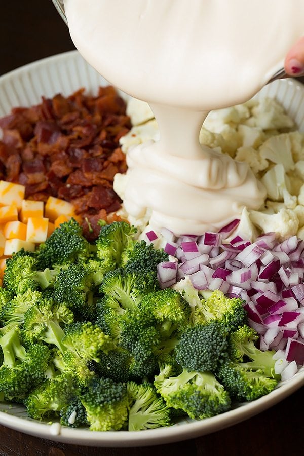 Broccoli and Cauliflower Salad will quickly become a staple recipes! It's always a crowd favorite, who knew people could love broccoli so much? | Broccoli Salad | Salad Side Dish | Salad With Bacon | #cookingclassy #broccoli #salad #bacon #cheese Broccoli and Cauliflower Salad ingredients in a white bowl with the dressing getting poured over the top