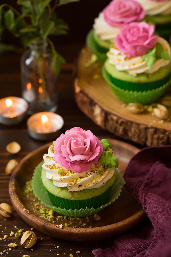 Pistachio Cupcakes One pistachio cupcake on a wooden dessert plate. Cupcake is light green with a white buttercream frosting and it's topped with a pink piped rose.