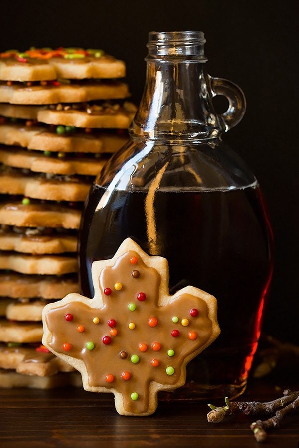Glazed Maple Shortbread Cookies Shortbread cookie flavored with pale cut into leaf shape and set in front of a jar of maple syrup, in the background is a large stack of the shortbread cookies.
