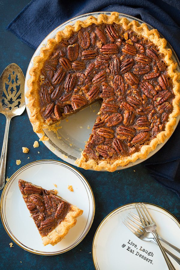 Pecan Pie Recipe Overhead image of whole pecan pie in pie dish and a slice to one side. Pie is resting on a blue surface.