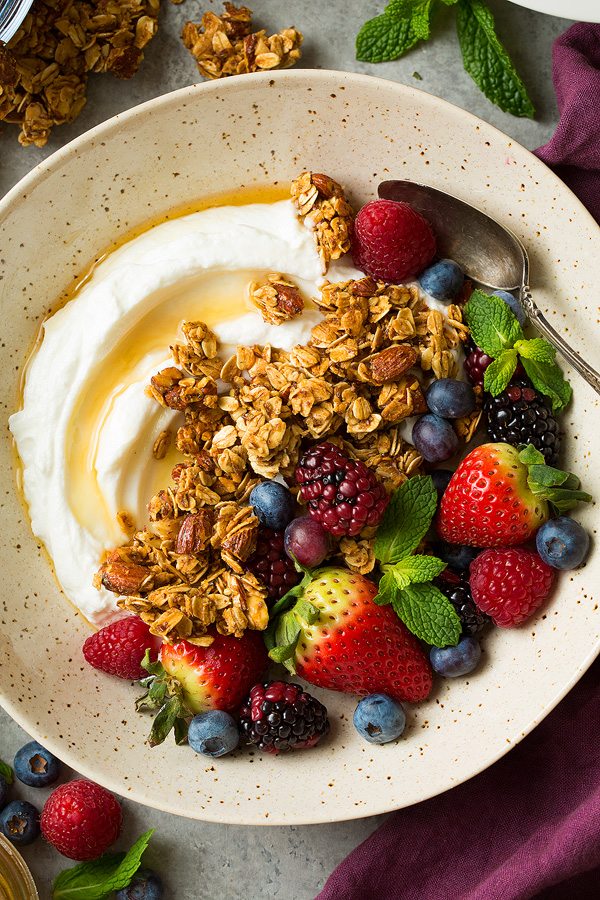 Honey Almond Granola Close up image of granola, fresh berries, yogurt and honey in a speckled beige bowl.