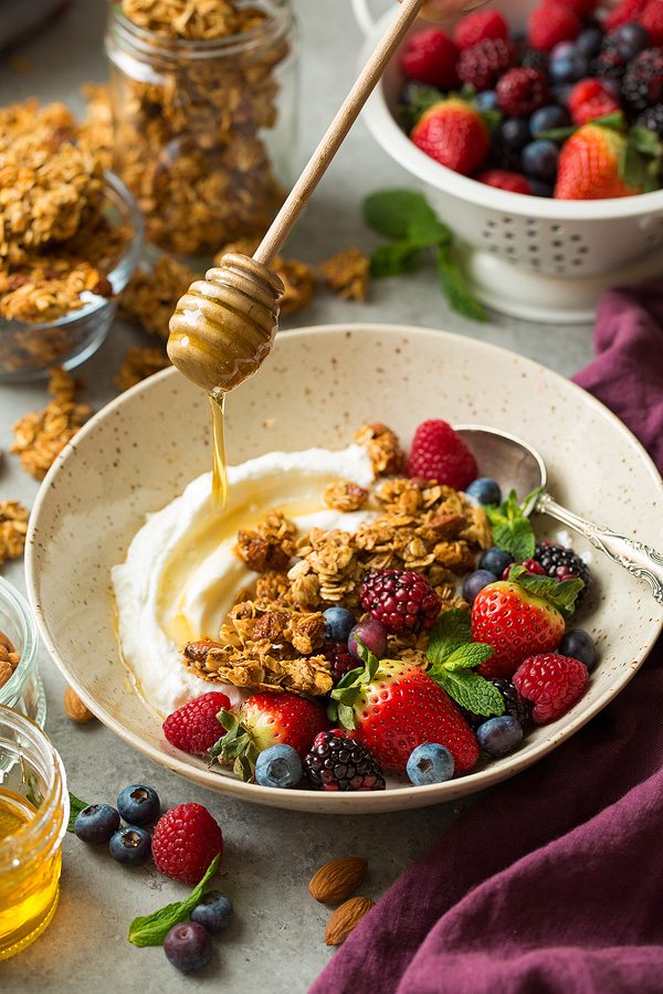 Honey Almond Granola Drizzling honey over granola, fruit and yogurt in a bowl.