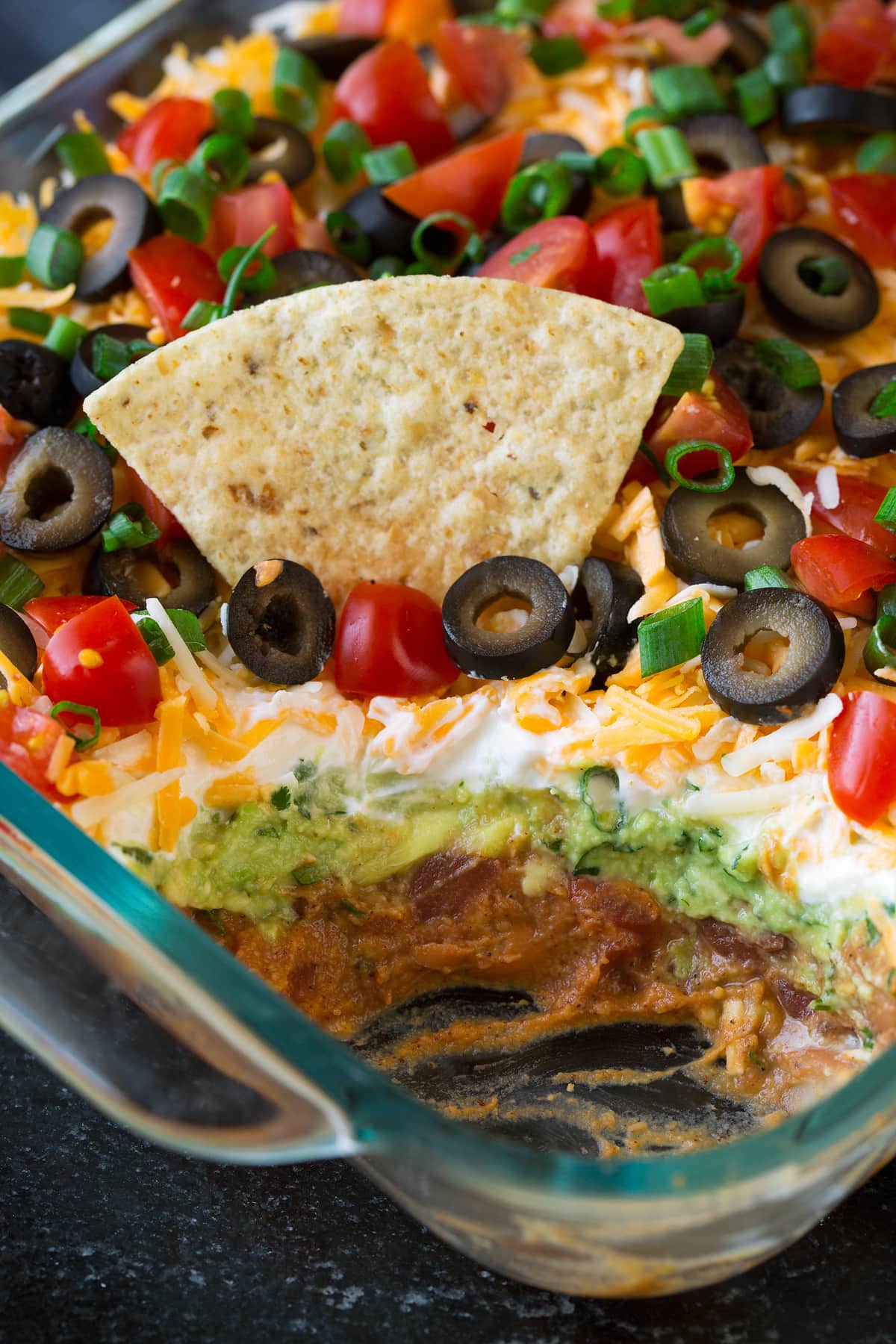 7 Layer Dip showing layer so of refried beans, guacamole, sour cream, cheese, olives, tomatoes and green onions.
