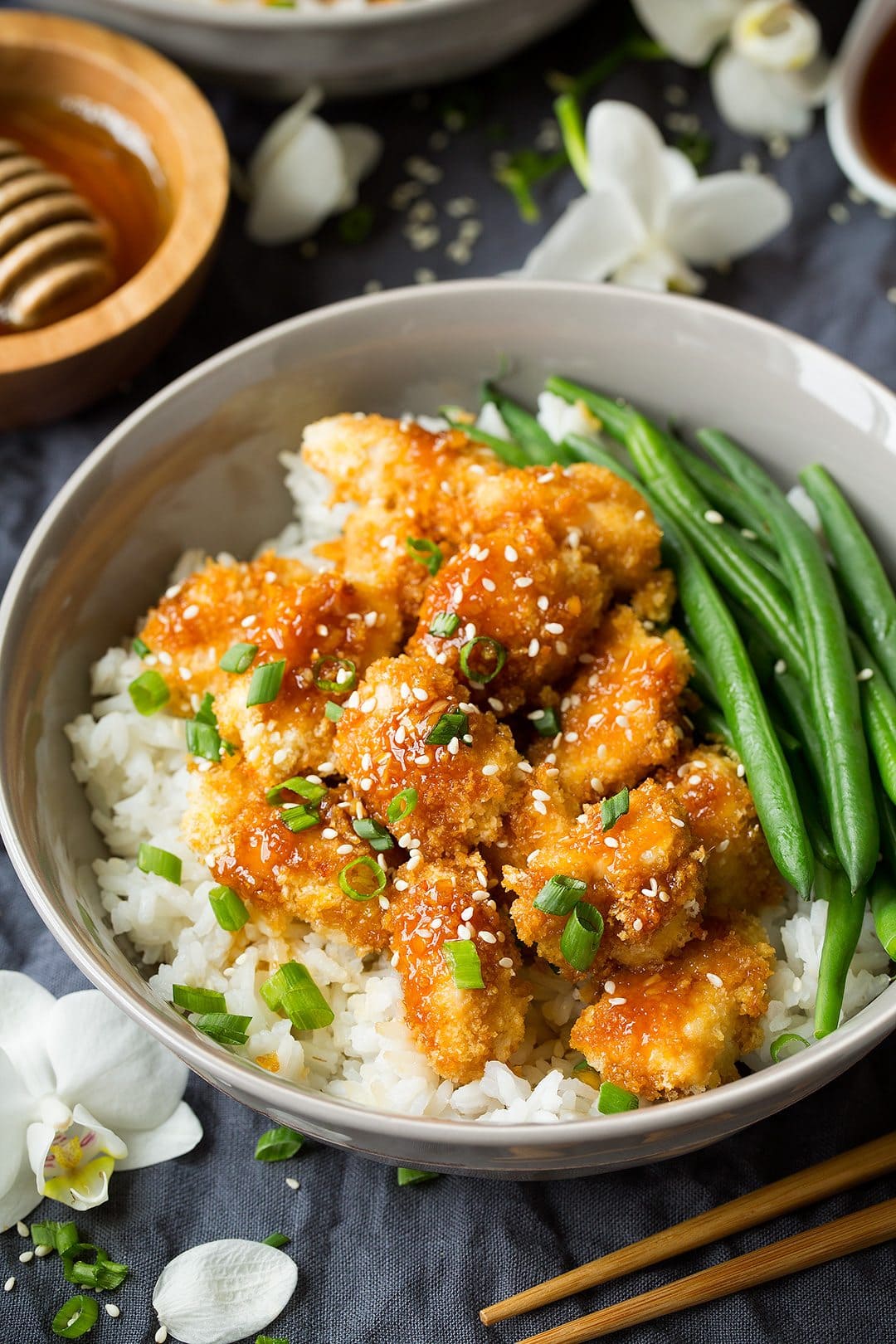 Honey Garlic Chicken Honey Garlic Chicken set over a layer of rice in a grey bowl with a side of green beans. Bowl sitting on a grey tablecloth and decorated with white orchids.