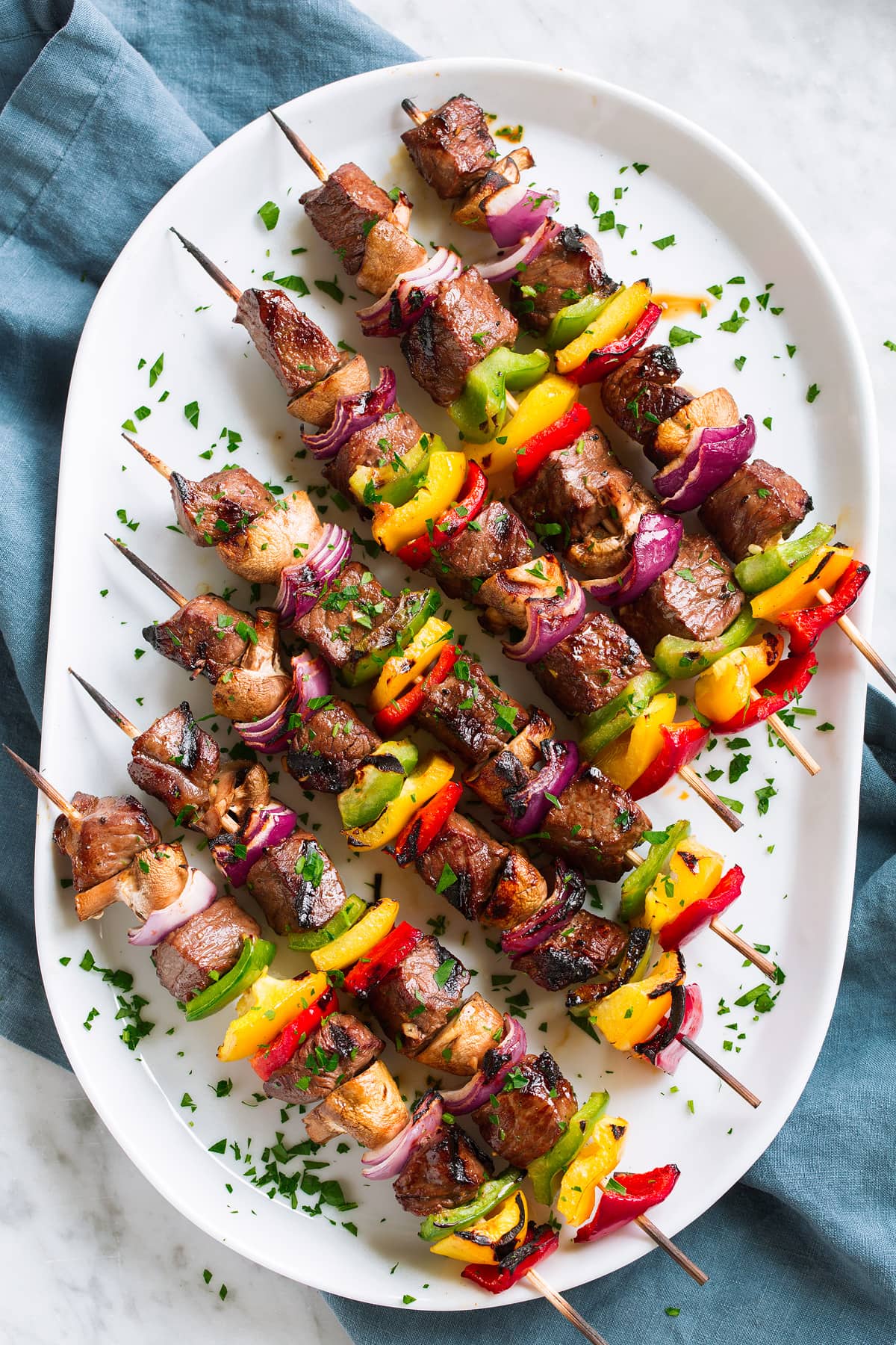 Steak Kebabs Seven steak kebabs on wooden skewers on a white platter set over a blue cloth on a marble table.