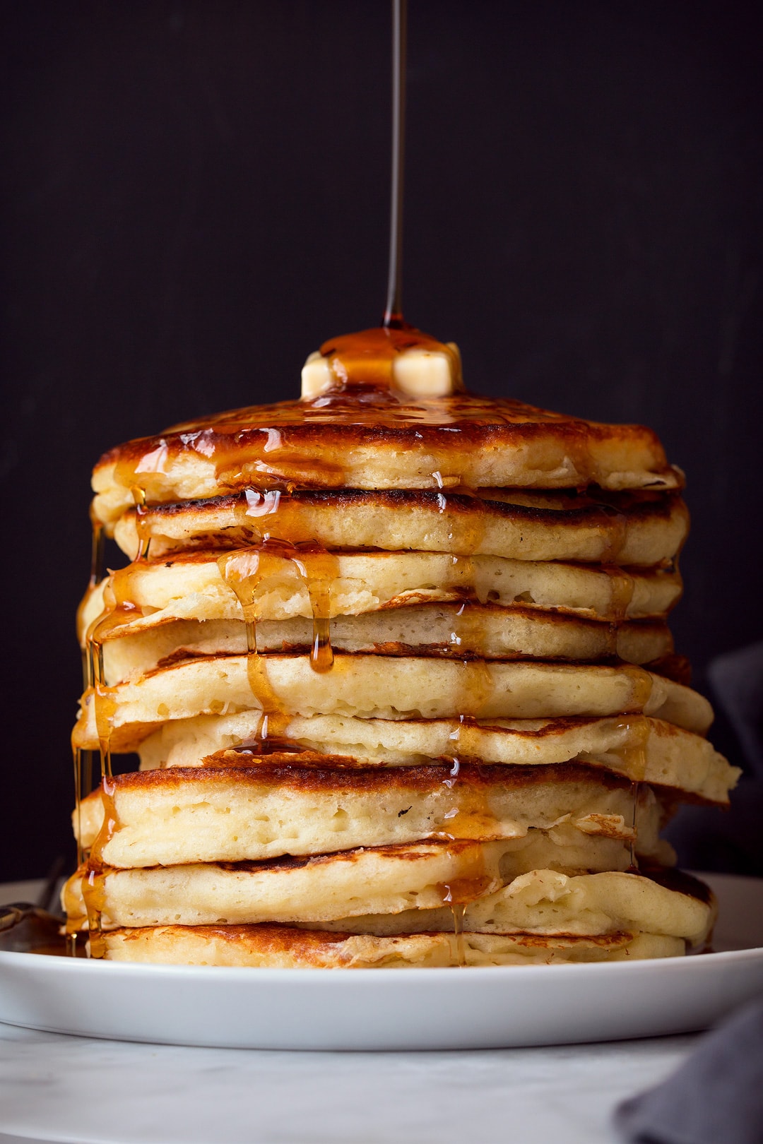 Buttermilk Pancakes Maple syrup being poured over a tall stack of pancakes with a dab of butter on top. Pancakes are sitting on a large white plate on a marble surface, background is black.