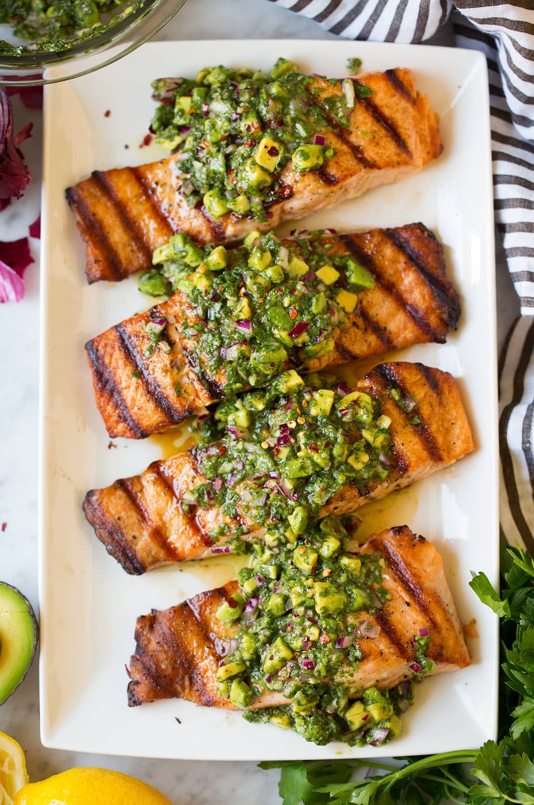 Four grilled salmon fillets on a plate topped with avocado chimichurri.