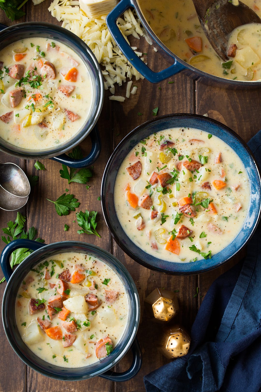 Three individual servings of Potato Soup with Sausage and White Cheddar shown here in a navy bowls over a brown wooden surface.