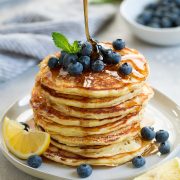 stack of eight lemon ricotta pancakes topped with fresh blueberries and mint maple syrup poured over pancakes lemon slices added on plate for garnish