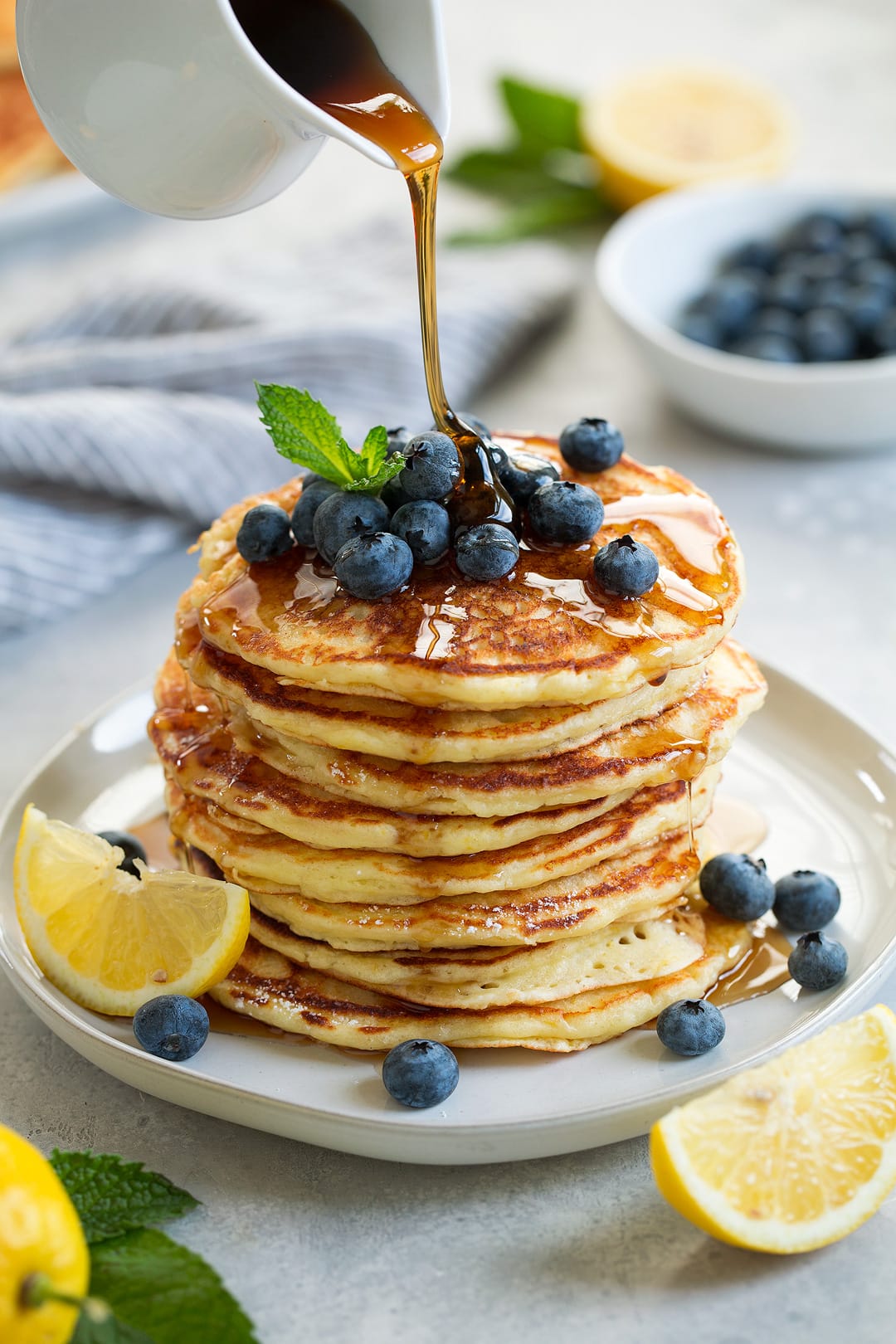 stack of eight lemon ricotta pancakes topped with fresh blueberries and mint maple syrup poured over pancakes lemon slices added on plate for garnish