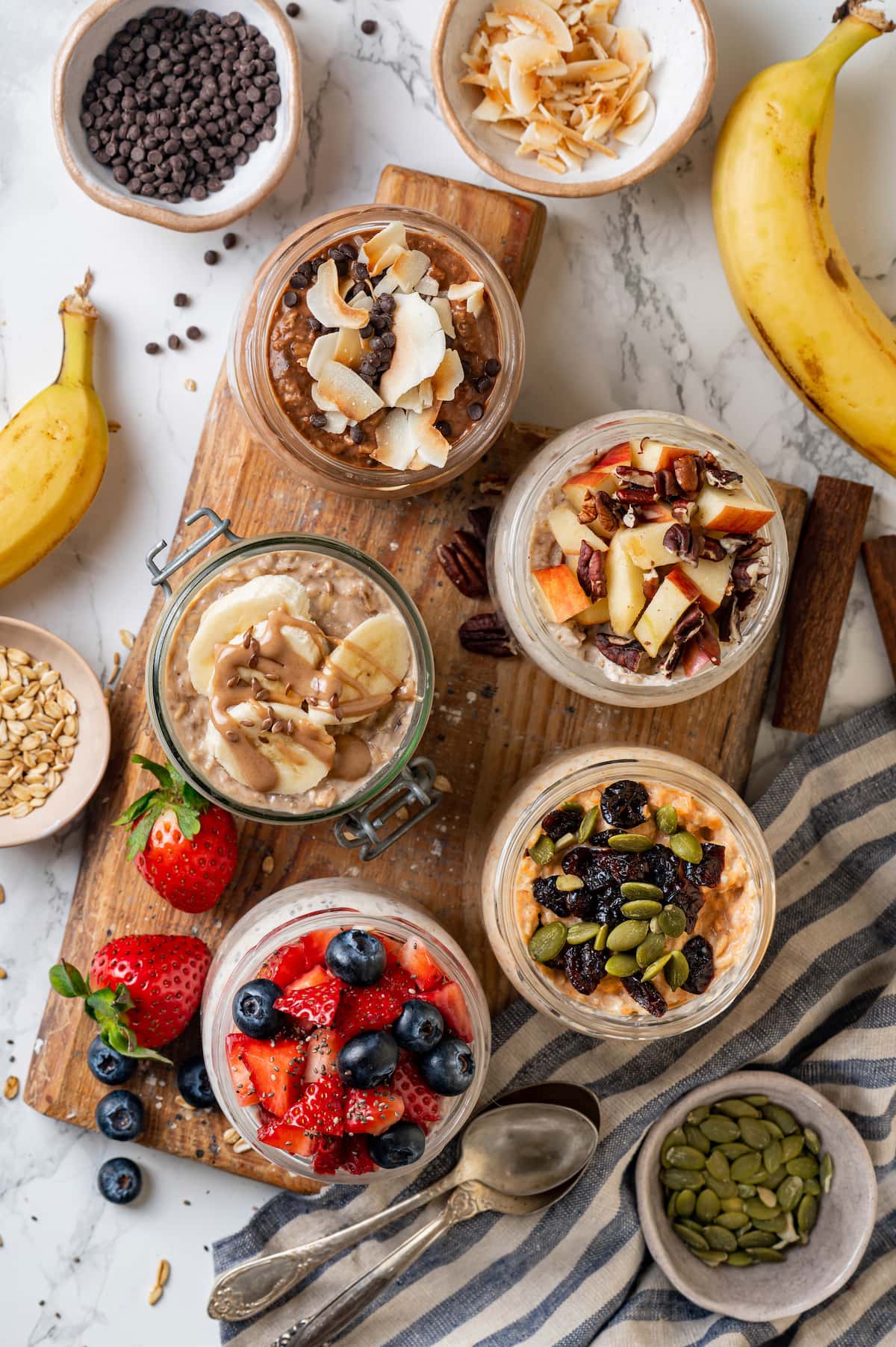 Learn how to make the best overnight oats that are rich, creamy, and perfect for breakfast on the go. Change it up with 5 amazing flavors! Overhead view of overnight oats in mason jars, five different ways, on a wooden platter surrounded by bowls of toppings and add-ins.