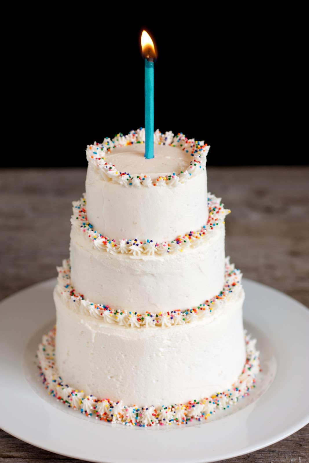 Buttercream Frosting Three tiered cake covered with the best vanilla buttercream frosting. Cake edges are garnished with multi colored sprinkles and there's one lit candle atop the cake.