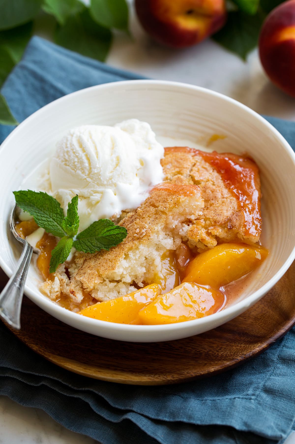 Peach Cobbler - the only peach cobbler recipe you'll ever need! I've never found better! Light and fluffy, buttery topping and perfectly sweet and juicy peach filling. #peach #cobbler #dessert #recipe Serving of homemade peach cobbler in a white serving bowl topped with vanilla ice cream and mint.