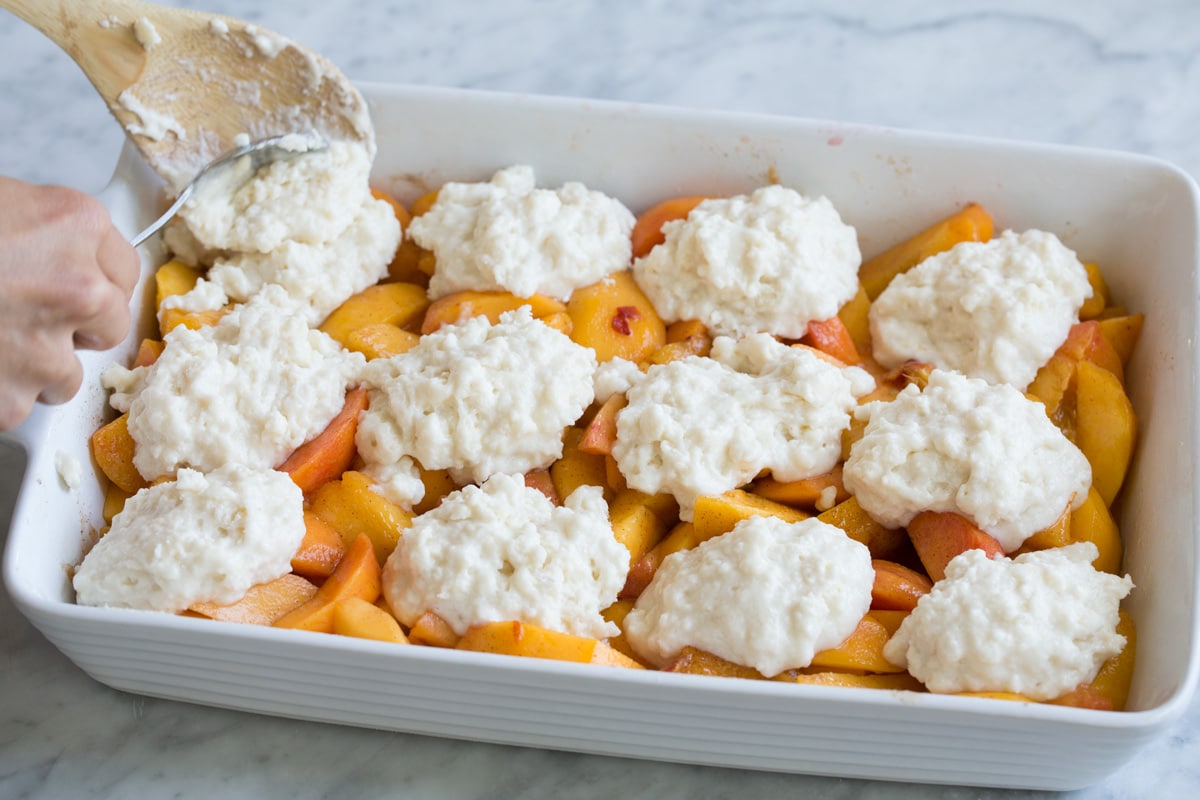 Peach Cobbler - the only peach cobbler recipe you'll ever need! I've never found better! Light and fluffy, buttery topping and perfectly sweet and juicy peach filling. #peach #cobbler #dessert #recipe dolloping cobbler topping over peach layer in baking dish.