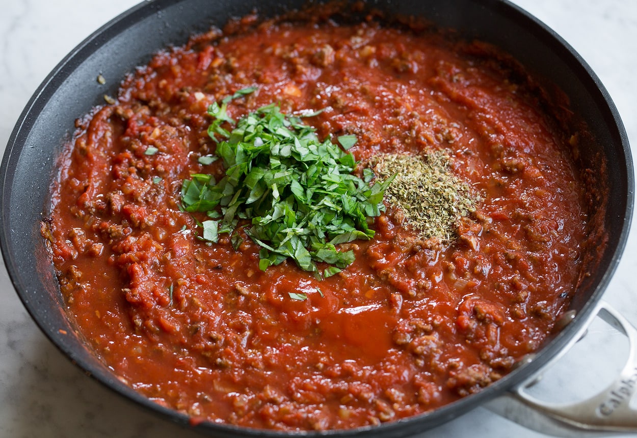 Homemade Spaghetti Sauce Adding basil and oregano to beefy tomato spaghetti sauce in pan.