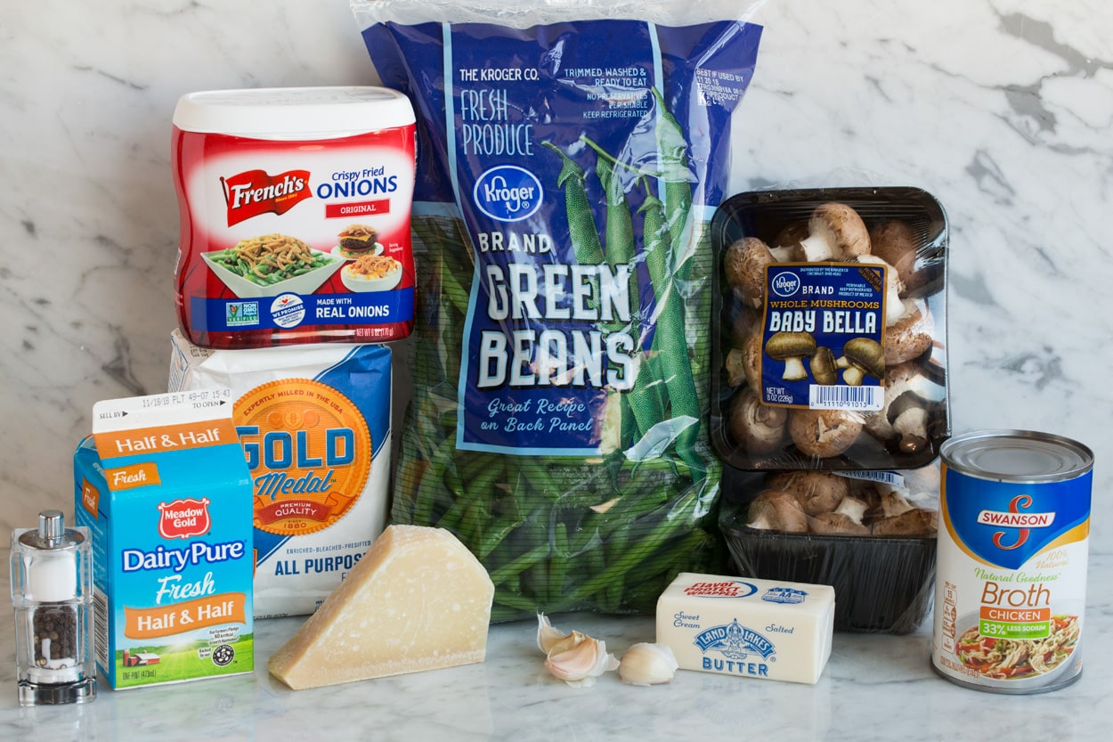 Green Bean Casserole Ingredients for Green Bean Casserole shown here on a white marble surface. They include fresh green beans, mushrooms, French's fried onions, flour, butter, half and half, chicken broth, parmesan, and garlic.