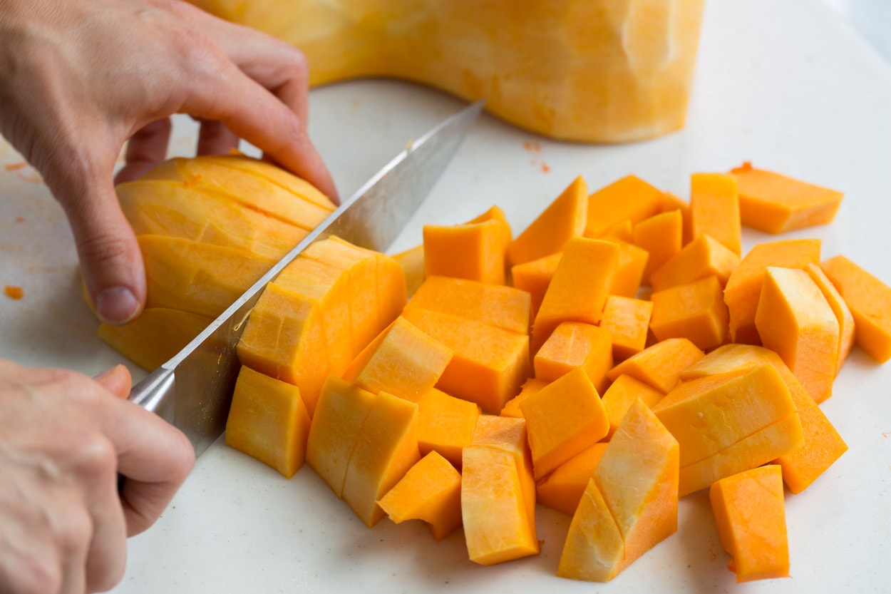 Roasted Butternut Squash with Garlic and Herbs Showing how to cut a whole butternut squash into cubes on a cutting board with a knife.