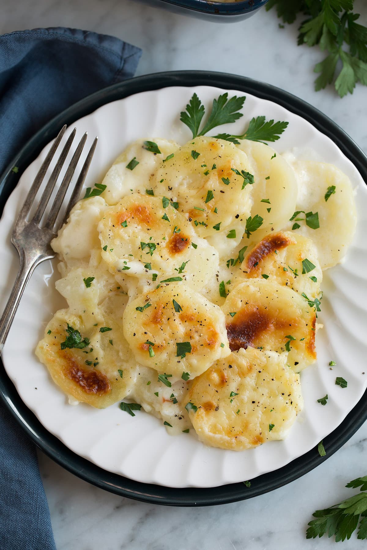 Scalloped Potatoes Scalloped Potatoes on a white single serving dish set over a dark blue plate that's resting on a marble surface.
