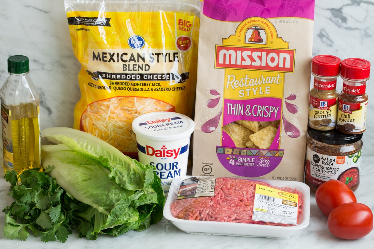 Taco Salad - so easy and flavorful! Image showing ingredients needed for taco salad including ground beef, olive oil, spices, bottled salsa, chips, cheese, sour cream, tomatoes.