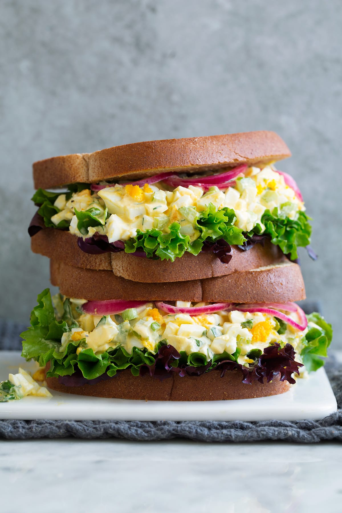 Egg Salad Stack of two Egg Salad Sandwiches on a white serving platter sitting on marble with a grey background.