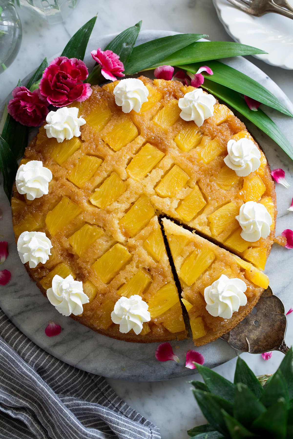 Pineapple Upside Down Cake Overhead image of Pineapple Upside Down Cake showing rows of pineapple chunks. Cake is decorated with swirls of whipped cream. Along the edge of the cake are pink flowers and pineapple leaves for decoration.