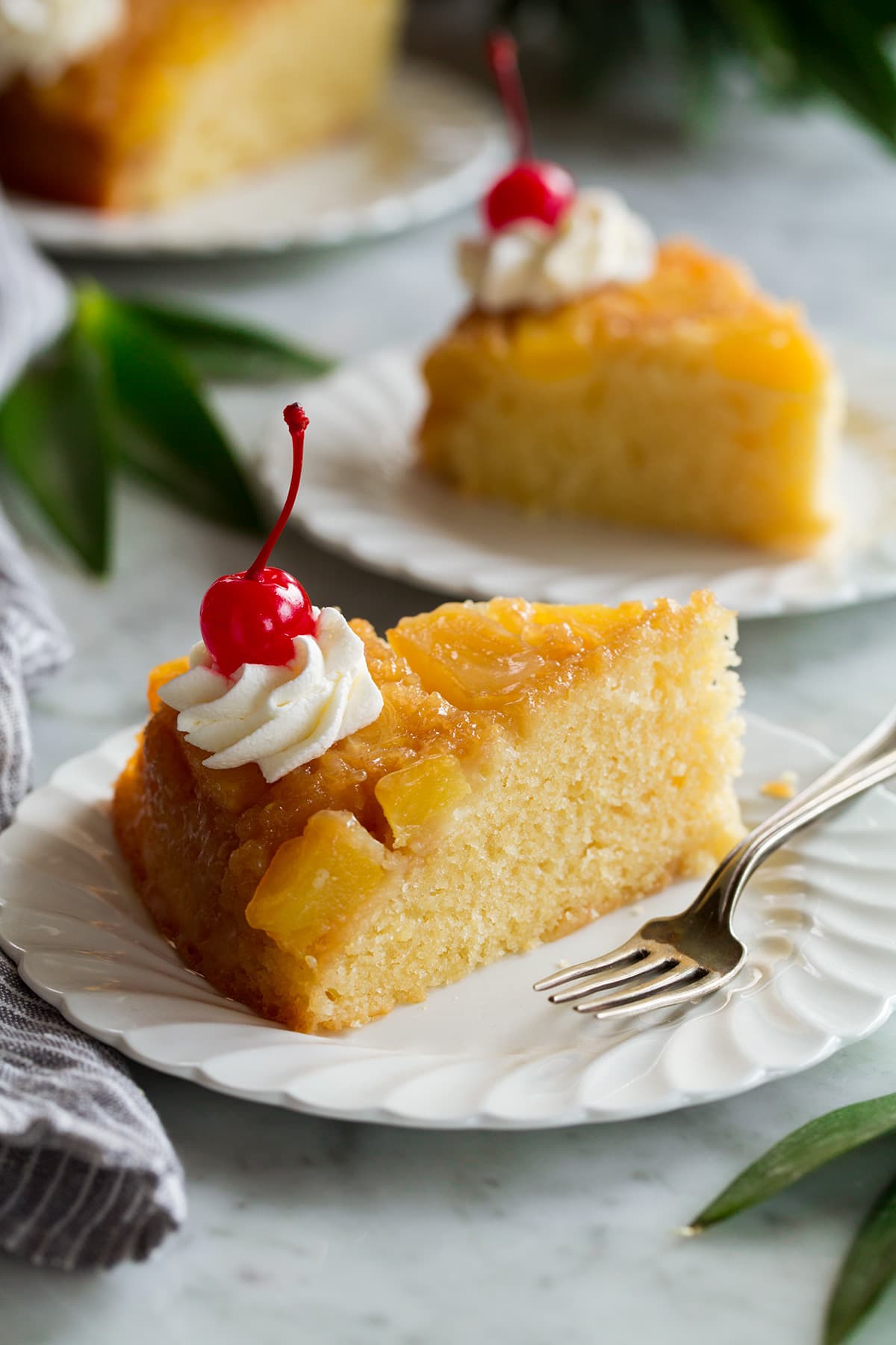 Slice of Pineapple Upside Down Cake on a white serving plate. Cake is topped with fresh pineapple slices and decorated with whipped cream and a maraschino cherry.