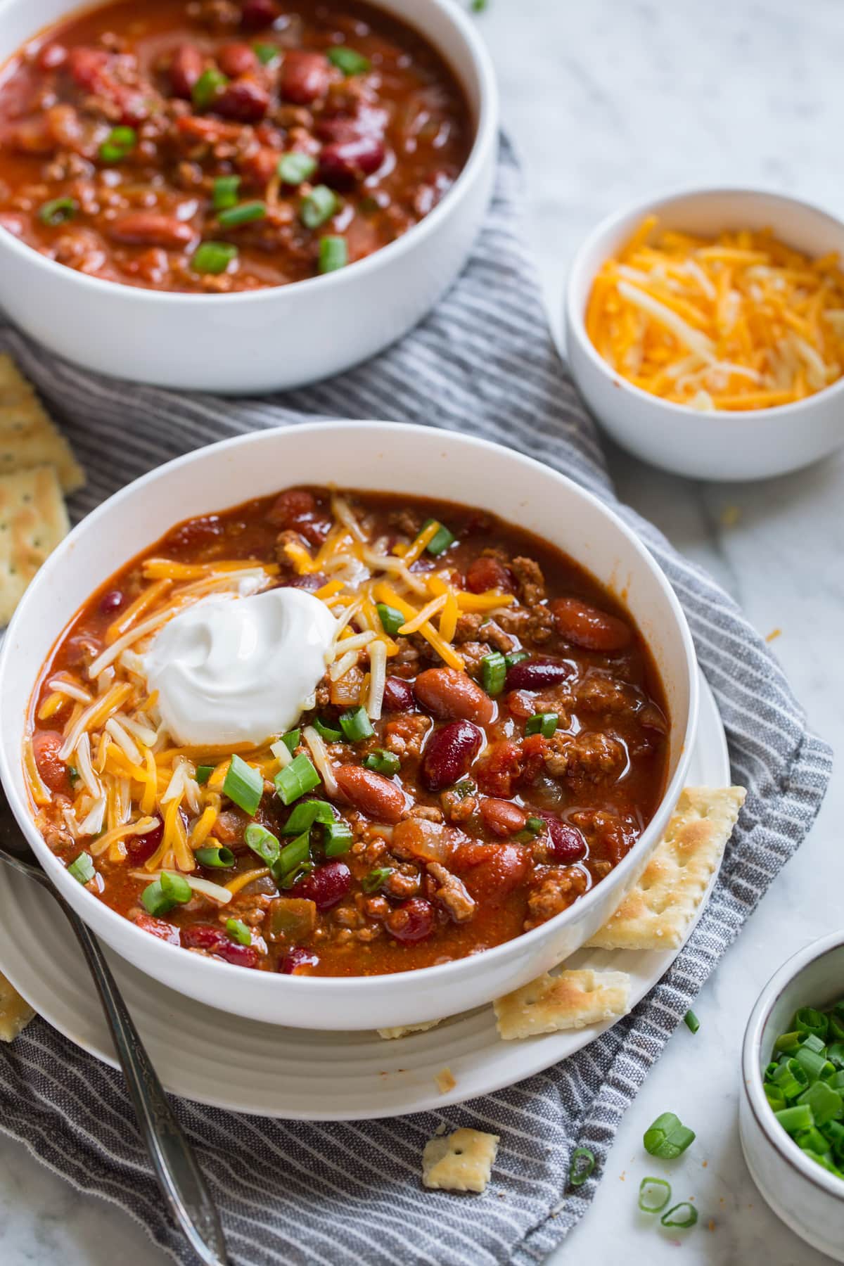 BEST Ever Chili Recipe! Two servings of chili in white bowls set over a marble surface. Servings topped with cheddar, sour cream and green onions.