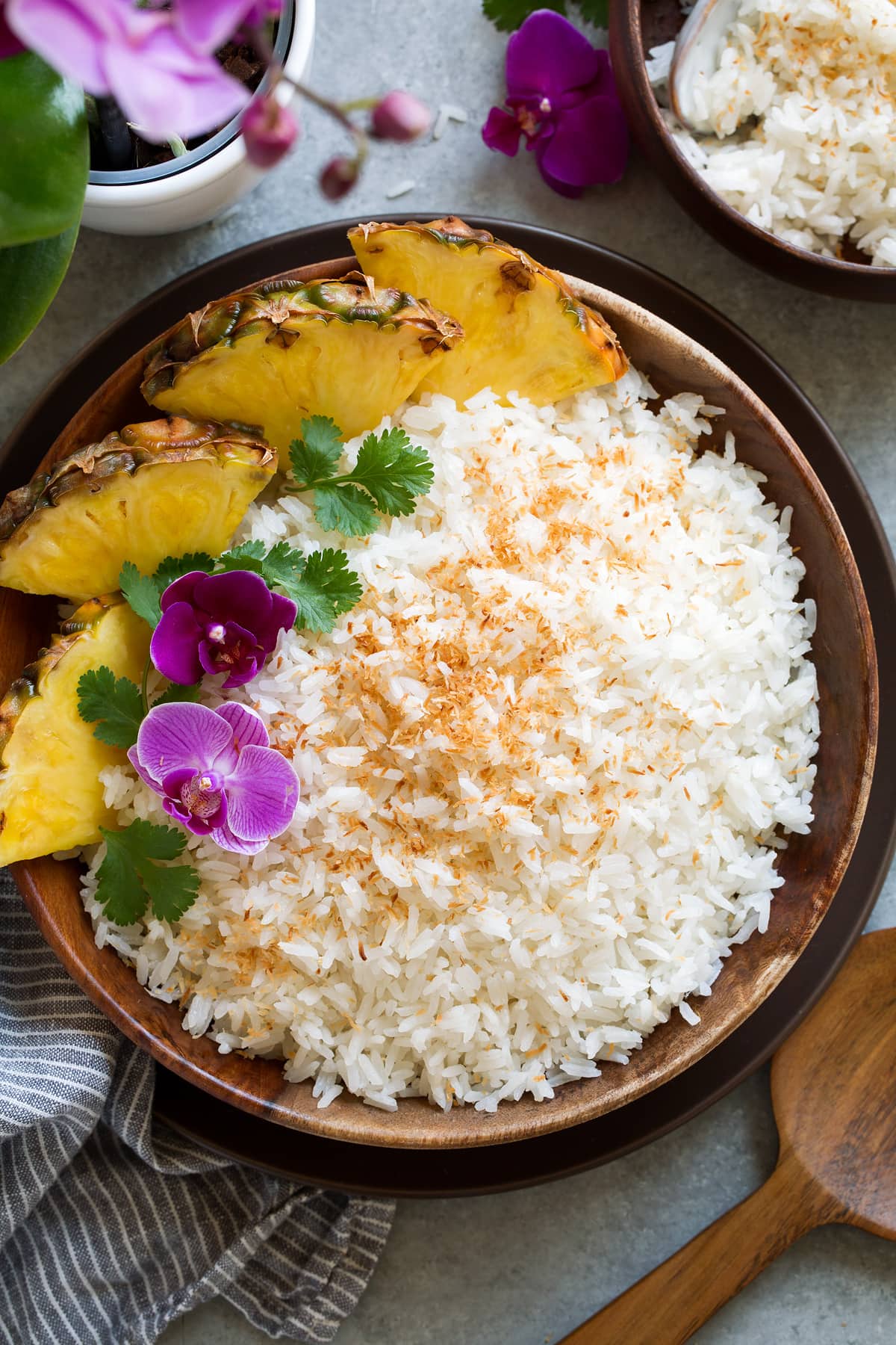 Coconut Rice Wooden bowl full of Coconut Rice topped with flowers, cilantro and a side of fresh pineapple.